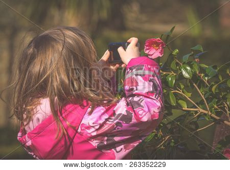 Cute Little Caucasian Girl Taking Photos Of Flowers In The Park In Spring