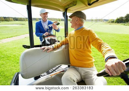 Senior man sitting in golf car while talking to his buddy preparing golf clubs behind