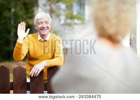 Cheerful and friendly mature man waving hand to his neighbour while standing by fence