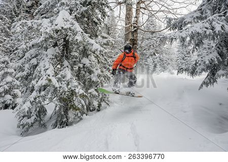 Man Riding Down The Hill On The Green Snowboard Near The Trees In The Mountain Tourist Resort In God
