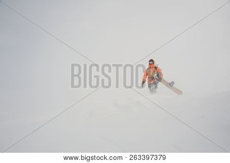 Man In The Orange Sportwear Walking With The Snowboard In Hands In The Mountain Up The Hill In The P