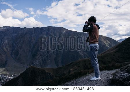 Young Curly Traveler Taking Photo With Camera In Kazbegi. Beautiful View Of Kazbegi In Autumn