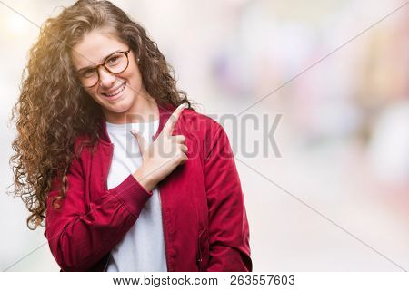 Beautiful brunette curly hair young girl wearing jacket and glasses over isolated background cheerful with a smile of face pointing with hand and finger up to the side with happy and natural expressio
