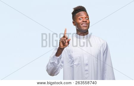 Young arabic african man wearing traditional djellaba over isolated background pointing finger up with successful idea. Exited and happy. Number one.