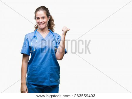 Young brunette doctor girl wearing nurse or surgeon uniform over isolated background smiling with happy face looking and pointing to the side with thumb up.