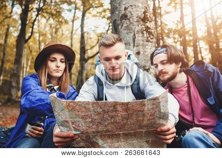 Group Of Young Creative Tourists With Bacpacks Reading Map Around The Trees And Make Up The Route, T