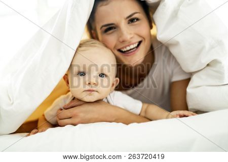 Portrait Of Young Mother With Infant Baby Girl Liying On The Bed Covered With A White Blanket
