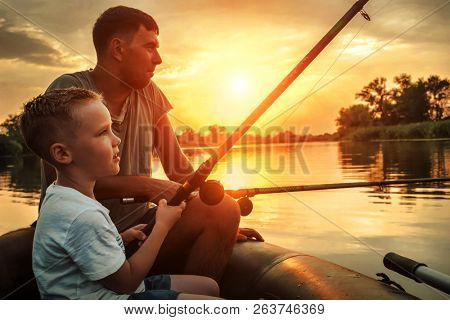 Happy Father and Son together fishing from a boat at sunset time in summer day under beautiful sky on the lake.