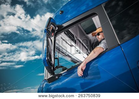 Heavy Load Cargo Delivery. Caucasian Truck Driver Inside Semi Tractor Cabin And The Blue Sky. Transp