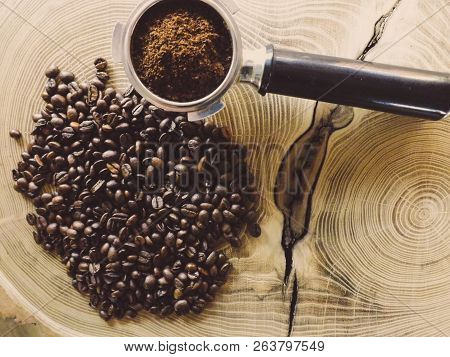 Close Up Of A Metal Portafilter Filled With Coffee Powder And Coffee Beans Around On Wooden Table