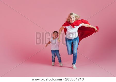 Super Mom And Her Son Run Forward. Fun Family, A Young Blond Woman In A Red Cape As Superhero, Pink 