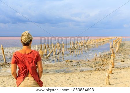 Young Woman On The Coast The Salty Lake With Pink Water And The Beach From Salt. Old Logs Pier On La