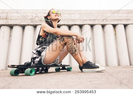 Young Girl With Tattoo And Dreadlocks Sitting On A Longboard On The Background Of Concrete Structure