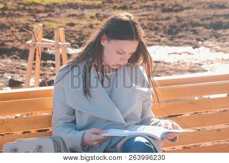 Young Beautiful Brunette Woman Student Sits On The Bench In Spring Park, Reads A Book.