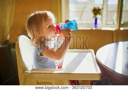 Adorable Toddler Girl Sitting In High Chair At Home Or At Restaurant And Drinking Water From Sippy C