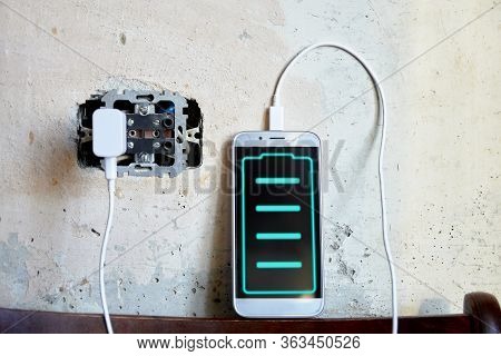 Phone Is Charging Against The Background Of A Concrete Wall In A Wall Outlet. Level Indicator On The