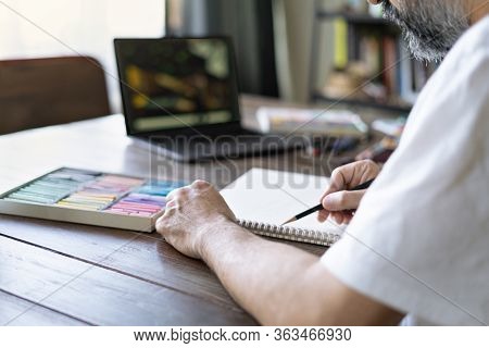 Man Painting With A Pastel Crayon Chalk And Color Pencils On Sketch Book, In Front Of Laptop Learnin