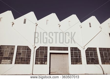Old-fashioned Storage Industrial Building With Closed Roller Door And Saw-toothed Roof Profile.