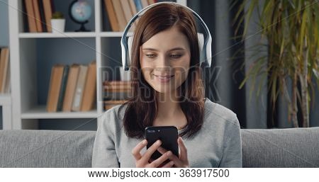 Headshot Of Glad Young Female Using Smartphone And Wireless Headphones To Divert Herself At Home