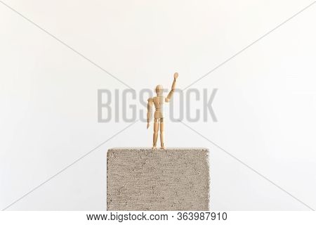 Wooden Human Figure Standing Isolated On A Concrete Block, Waving With A Raised Hand. Symbol Of Self