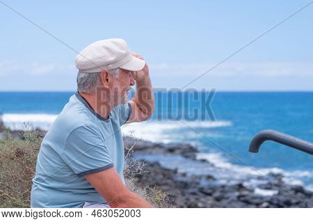 Smiling Caucasian Bearded Senior Man With Hat Sitting At The Beach Looking At The Horizon While Enjo