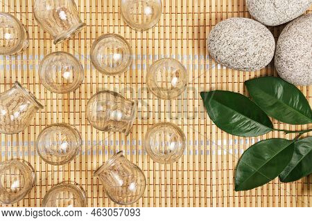 Glass Cups For Cupping Therapy On A Bamboo Mat With Stones And A Green Twig, Flat Lay Composition.