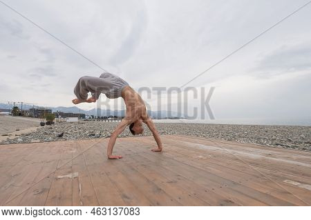 Shirtless Caucasian Man Doing Backflip On Pebble Beach.