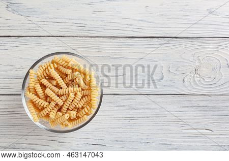Uncooked Fusilli Pasta In Glass Bowl On White Wooden Background With Copyspace