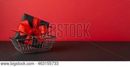 Black Friday Wholesale Concept. Photo Of Black Gift Box With Red Ribbon Bow In Shopping Cart On Blac