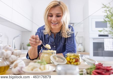 Sniling Blond Woman Picks A Teaspoon Of Crushed Garlic From A Jar In The Kitchen.