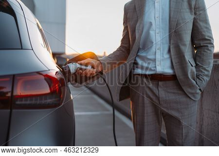 Businessman, Holding Power Supply And Charging His Electric Car During Sunset. Concept Of Ecology Tr