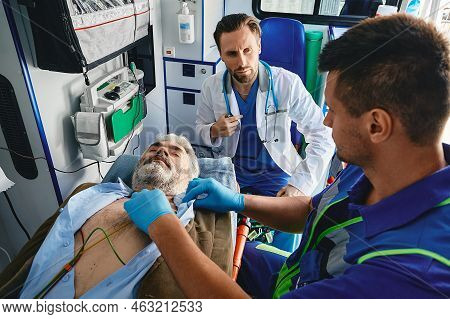 Cardiac Patient In Ambulance. Paramedics Performing Ecg On Male Vicitm Lying On Stretcher With Elect