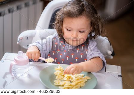 Portrait Caucasian Baby Girl About 2 Years Old In Bib Eating Pasta From Plate Sitting High Chair, Se