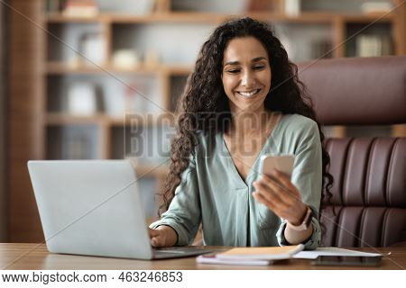 Happy Middle Eastern Woman Sitting At Workdesk, Using Smartphone