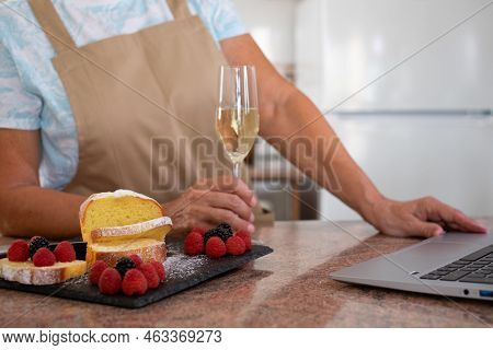 Senior Woman Holding A Glass Of Wine While Browsing On Laptop. Homemade Plumcake Decorated With Berr