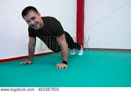 Portrait Of A Strong Determined Young Man Doing Push-ups And Looking At Camera Smiling. He Is Dresse