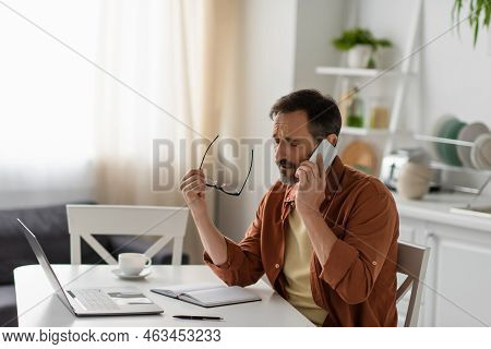 Frustrated Man With Closed Eyes Holding Eyeglasses And Talking On Mobile Phone Near Laptop.