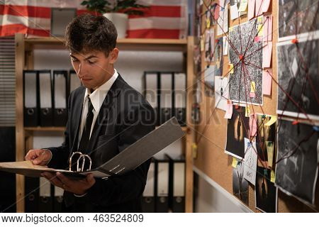 Concentrated Young Detective In Black Suit Reading Dossier In Dark Office. Investigator Working With