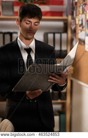 Concentrated Young Detective In Black Suit Reading Dossier In Dark Office. Investigator Working With