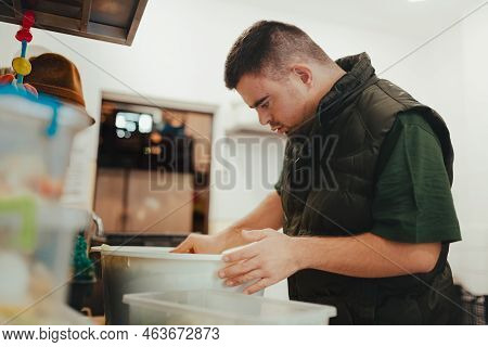 Man With Down Syndrome Working In Kitchen In Zoo, Prepairing Food For Animals.concept Of Integration
