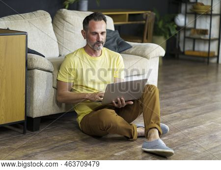 Attractive Mature Man Dressed In Yellow T-shirt Sitting On The Floor With Laptop Computer Browsing I