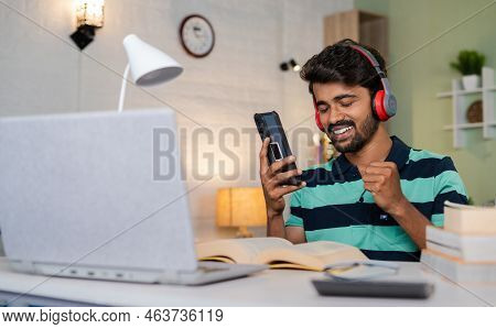 Enjoying Young Man While Listening Songs On Mobile Phone With Wirless Headphones In Front Of Laptop 