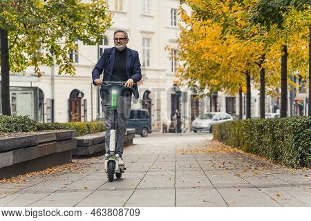 Middle-aged Gray-haired Man In Formal Clothes Riding Rental Electric Scooter To Work Through City Ce