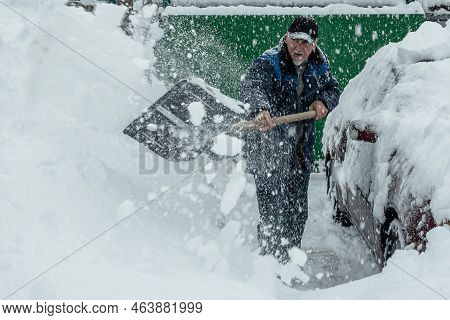 A Man Is Shoveling Snow From The Area Near The House. Nearby Is A Car Covered With Snow. It Is Snowi