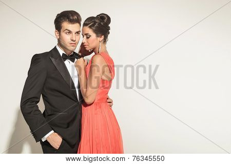 Picture of a young elegant couple posing, the man is looking away with one hand in his pocket, while his woman si embracing him.
