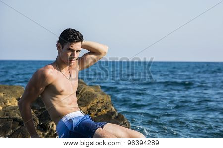 Young shirtless athletic man sitting on rock by sea