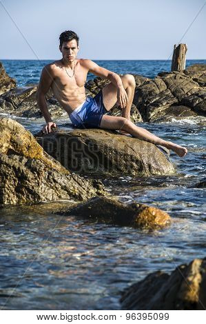 Young shirtless athletic man sitting on rock by sea