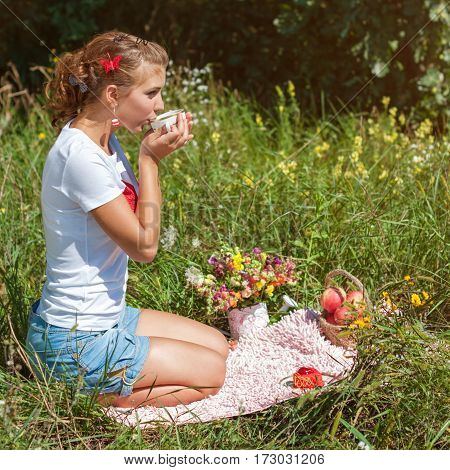 Beautiful young woman having picnic on sunny spring day in park drinking tea from the cup.