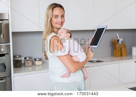 Portrait of mother using digital tablet while carrying her baby in kitchen at home