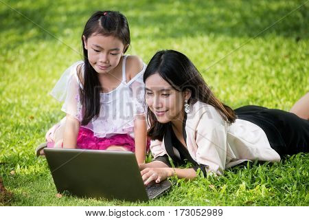 Close up portrait of little asian girl with braids and mother with laptop. Kid typing on laptop against green background outdoors.